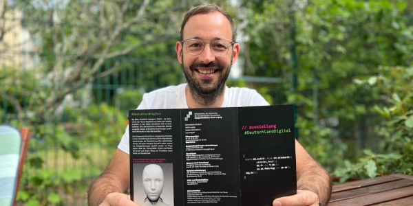 Daniel hält einen Flyer der Ausstellung #DeutschlandDigital in die Kamera.